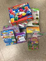 Top view photo showing various untidy stacked craft and sticker kits on a beige floor including RoseArt Funny Fingers finger paints, Wiggly Pets kit, and multiple Melissa & Doug sticker sets.