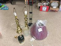 View of two brass lamps with harp frames and bulbs, a large maroon lamp shade sealed in plastic, white frosted glass shade, two black metal lamps, and a small dark glass lamp base with gold accent on tile floor