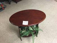 Front view of the wooden oval side table showing the reddish-brown surface, lower shelf with three faux green plants.