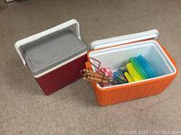 Two coolers on floor, one red and one orange containing colored plates and utensils with wooden handles.