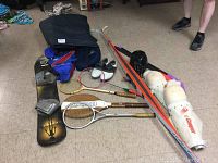 Wide view of sports lot showing skis, snowboard, rackets, skates, pads, and bag on carpeted floor with a person's legs visible.