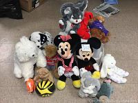 Photo showing a collection of 12 assorted stuffed animals including Disney Mickey and Minnie Mouse, multiple plush dogs, a teddy bear, and various other small plush toys on a floor.