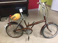 Full side view of the brown Raleigh Folder folding bicycle showing the frame, wheels, chain guard, seat, and front and rear fenders, with a yellow helmet attached on the rear rack.