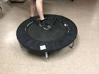 Top angle view of small exercise trampoline with black mat and black padding showing six silver metal legs and a person standing on it with black sneakers.