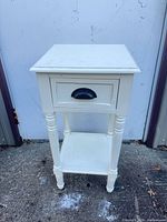 Front view of cream-painted wood side table showing drawer with black cup pull and lower shelf