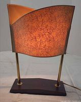 Front view of lit lamp showing curved shade, brass posts, wood base.