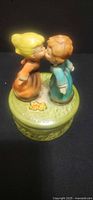 Front view of ceramic music box showing boy and girl figurines kissing on green base
