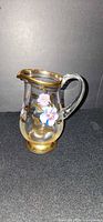 Side view of clear glass creamer showing floral enamel and gilded rim and base