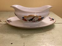 Side view of porcelain gravy boat attached to underplate showing bird and floral decoration and gold handle