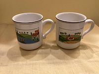 Front view of both Design Naif mugs showing village scene panels