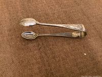 Full view of silver-plated sugar tongs showing ornate handles and spoon/scallop ends