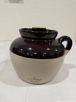 Front view of cream and brown stoneware bean pot showing handle, lid, and blue stamp