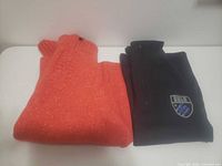 Folded orange knit sweater and black Bula fleece pullover displayed side by side