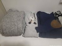 Three folded tops: light-grey chunky sweater, light-grey toggle cardigan, navy sweater