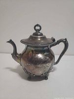 Front view of ornate silver-plated teapot showing body, spout, handle, lid and feet
