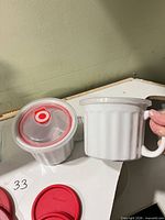 Round glass bowl with red vented lid alongside white ribbed ceramic mug; extra red lids visible