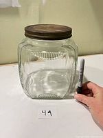 Square clear glass jar with decorative pattern and metal lid beside Sharpie for size reference