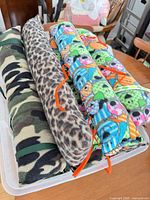 Top view of four fleece seat belt pillows in storage tub showing camouflage, leopard, leopard variant and cartoon prints with ribbon ties