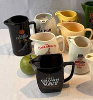 Group of ceramic advertising pitchers including Clan Campbell, VAT 69, The Famous Grouse, Grant’s, Beefeater Gin, Sykes Crown VAT plus apple for scale