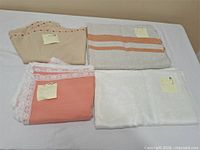 Four folded tablecloths on table: beige with stitched edge, off-white with orange stripes, coral with lace, white plain