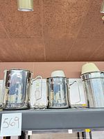 Three stainless steel pitchers on shelf: two measuring cups, one grease catcher with lid