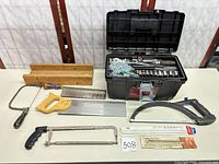 Overall lot layout: toolbox open with contents, wooden miter box, multiple saws and blades on table