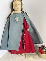 Front view of nun doll standing with cape, rosary, decorative box and paper slip visible