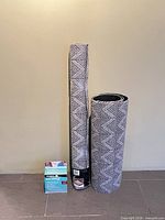 Two rolled utility mats and boxed rug gripper against wall