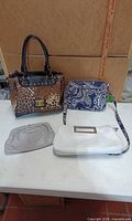 Four purses laid out on table: leopard satchel, paisley crossbody, white clutch, grey pouch