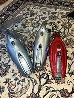 Three Dirt Devil handheld vacuums on rug: two silver, one red