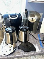 Both Keurig brewers, two carafes, drip trays and plastic accessory on countertop.