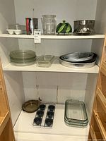 Shelved lot showing glass plates, baking dishes, cake pans, loaf pan, muffin pan, pie dish, steamer baskets, measuring pitcher, flour sifter, egg holder