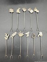 Full group of 11 sterling silver appetizer picks showing assorted figural tops and two-prong ends