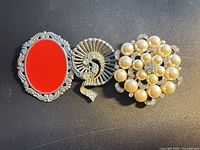 Three brooches side by side: red cabochon, rhinestone spiral, pearl cluster