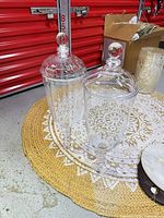Both glass apothecary jars standing on rug showing size, stems, etched designs, and lids with knobs