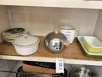 Shelf with decorative plates, white CorningWare casserole, chrome ice bucket, stack of petite blue cornflower casseroles, green and yellow Pyrex visible