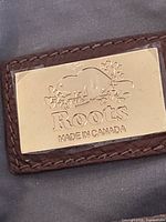 Gold-tone Roots beaver logo plaque on brown material