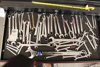 Top view of a selection of combination wrenches in various sizes; mostly chrome finish showing some wear and patina. Tape measure placed above for scale.