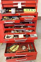 Red metal tool chest with multiple drawers open, showing various hand tools and supplies inside, including hammers, screwdrivers, and fasteners.