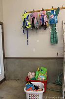 Four children's life jackets and a mermaid tail costume hanging on hooks above plastic bins containing assorted swim toys and a basketball on the concrete floor.