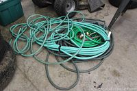 Five tangled and coiled garden hoses in multiple shades of green and blue on concrete floor near tires.