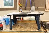 Side view of the Ryobi router table placed on a workbench, showing the black top surface and blue metal legs.