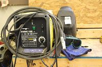 Overview of welder unit with cables, helmet, goggles and gloves arranged on bench