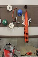 Photo showing wall mounted orange moving straps, blue and green hard hats, chalk line reels, measuring square, handsaw with orange handle, and shop light. Shop-Vac vacuum is on floor below.