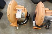 Two orange electric air pumps placed side by side on a concrete floor showing top and side views, labels visible on motors.