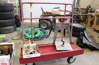 Red drywall trolley with metal frame and bottom tray. Background shows workshop environment with tires, coiled hose, and other tools.