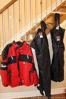 Photo showing the red and black Coldwave winter jacket hanging next to two pairs of black snow pants with suspenders on a stair railing.