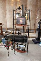 Full view of tall metal corner shelf stand with two reddish wooden semi-circular shelves and two small metal easels holding laminated prints, next to metal plant stand with leaf design