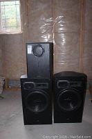 Three large black speakers in a room with unfinished walls. Two tall main speakers and one shorter subwoofer. Photos show wood grain texture and edges.