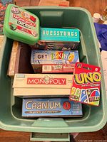 Green plastic tote containing Monopoly Diced, Guesstures, Get Packing, Scrabble, Monopoly, Cranium, Uno